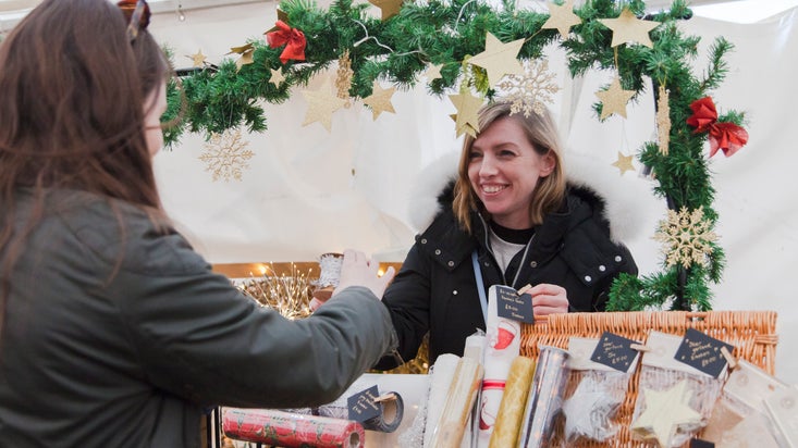 A woman smiles as she serves a customer from behind the counter of a Christmas Market stall. The stall is decorated with a festive garland and there is a wicker basket with wrapping paper and gift tags for sale.